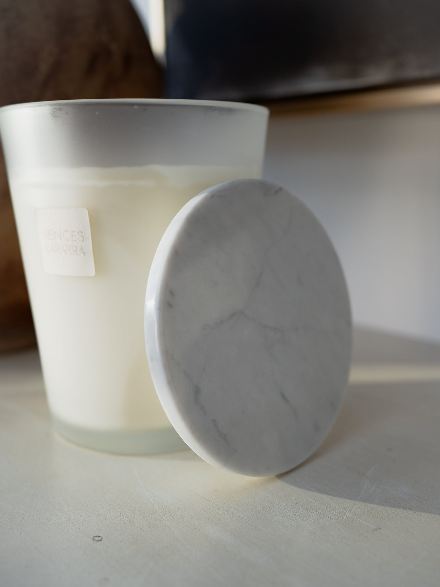 A frosted glass candle jar with white wax next to a round white marble LID on a light surface, with a softly blurred indoor background.