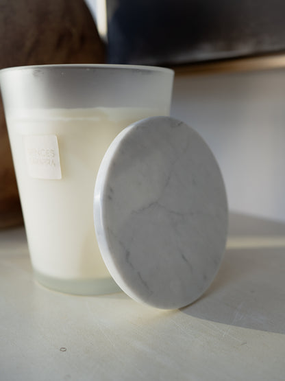 A frosted glass candle jar with white wax next to a round white marble LID on a light surface, with a softly blurred indoor background.