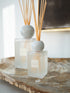 Two reed diffusers with frosted glass bottles, round white marble stoppers, and natural reed sticks, displayed on a rustic wooden tray.