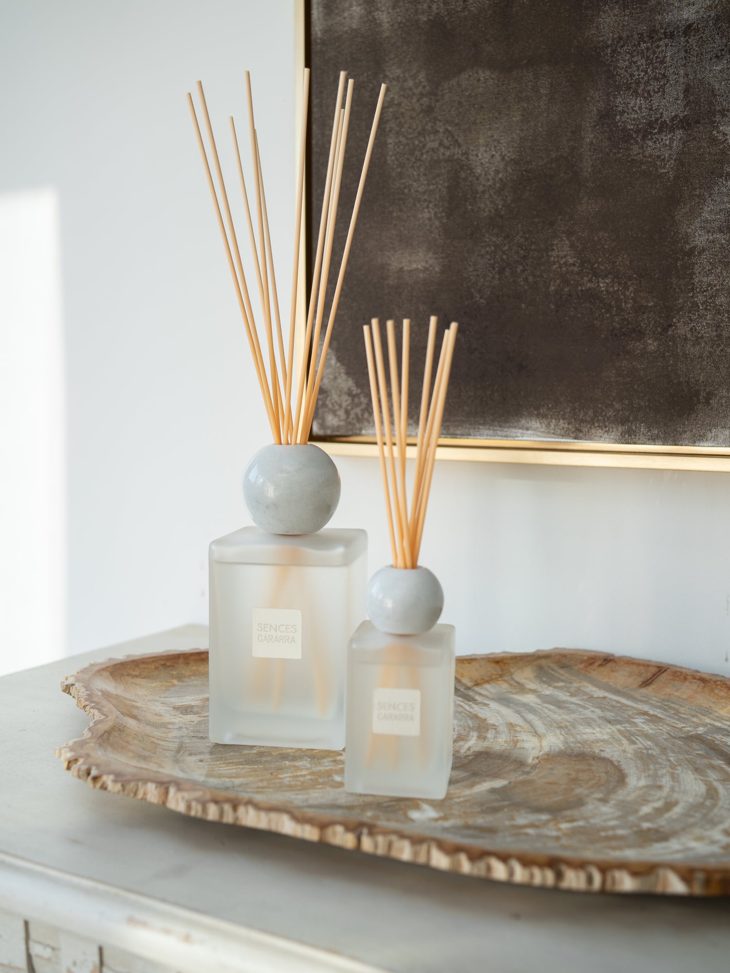 Two reed diffusers with frosted glass bottles, round gray marble, and natural reed sticks, styled on a rustic wood slice tray against a white wall and dark framed artwork.