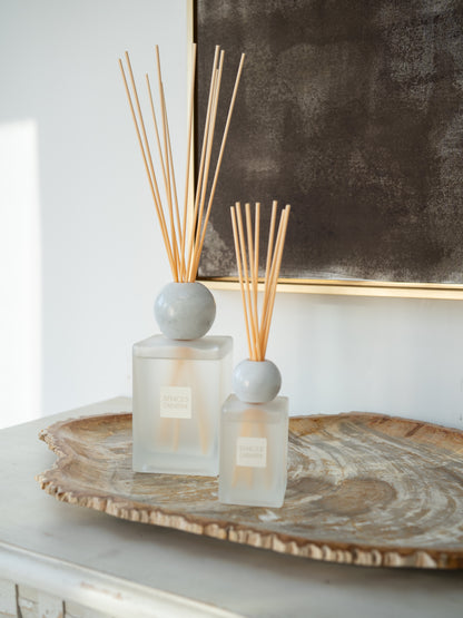 Two reed diffusers with frosted glass bottles, round gray marble, and natural reed sticks, styled on a rustic wood slice tray against a white wall and dark framed artwork.