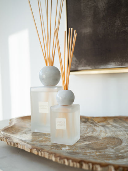 Two reed diffusers with frosted glass bottles and light gray spherical marble tops, placed on a rustic wood slice tray in a bright interior setting.