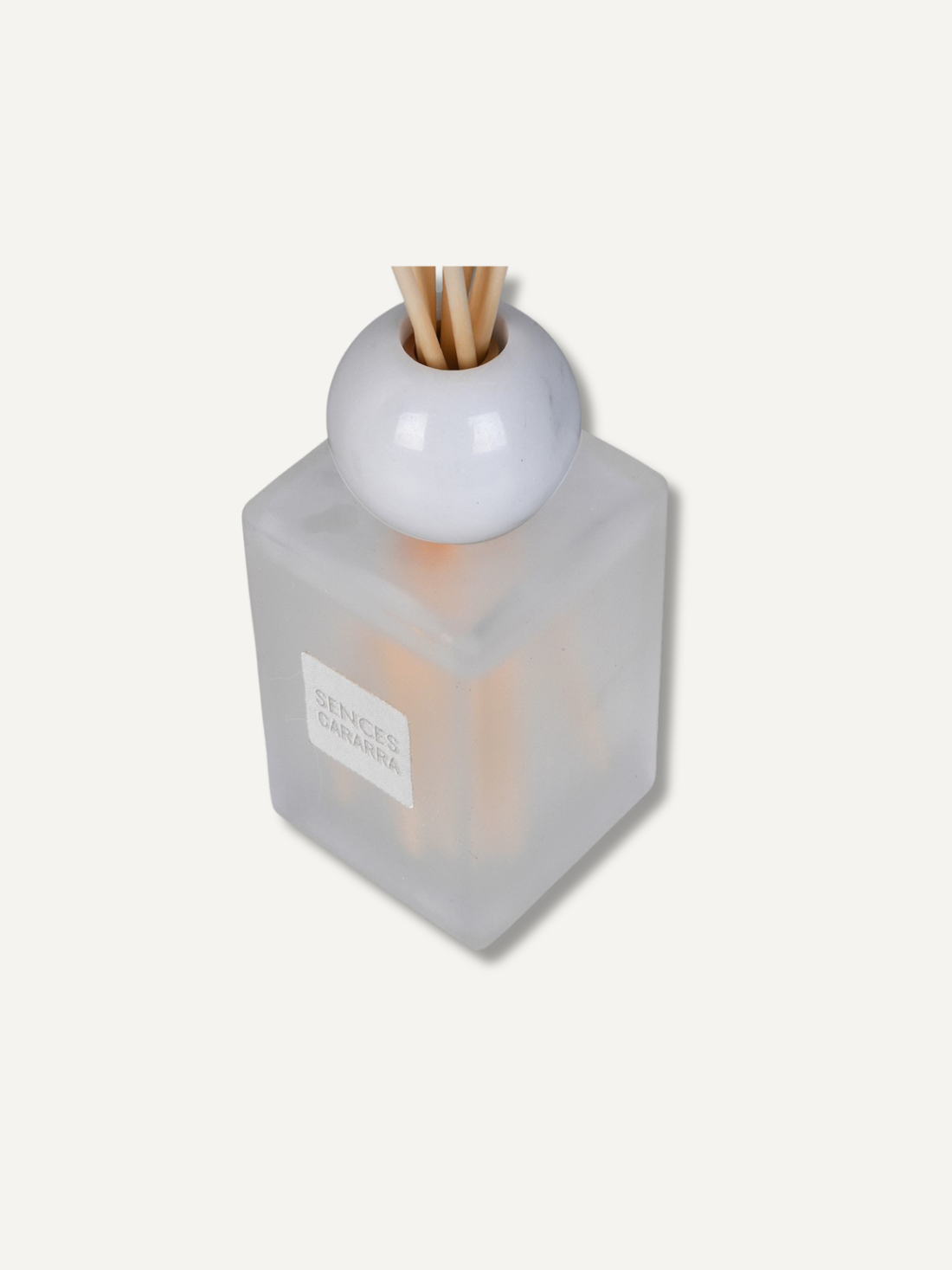 A reed diffuser bottle with a frosted translucent square glass body, a white spherical top, and natural reed sticks, shown on a plain light beige background.