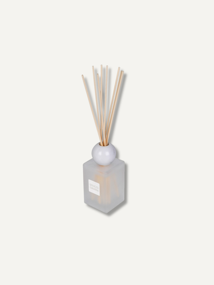 A reed diffuser with a frosted glass bottle, a white marble sphere, and natural beige reeds, shown on a solid off-white background.