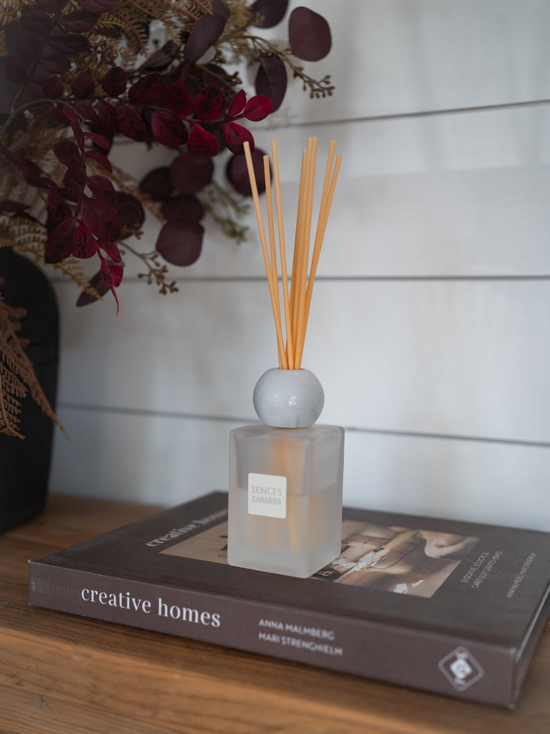 A reed diffuser bottle with light wood reeds sits on a book on a wooden table, with a white shiplap wall background and dried flowers in the upper left.
