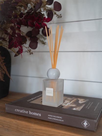 A reed diffuser bottle with light wood reeds sits on a book on a wooden table, with a white shiplap wall background and dried flowers in the upper left.