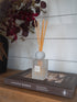 A reed diffuser bottle with light wood reeds sits on a book on a wooden table, with a white shiplap wall background and dried flowers in the upper left.