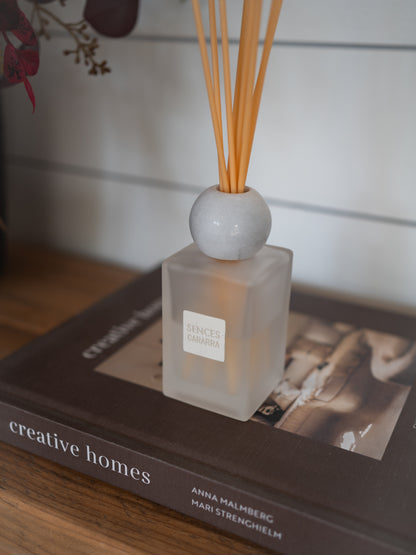 A reed diffuser bottle with a frosted square glass body, a round white marble-like top, and natural reed sticks, placed on top of a book in a home interior setting.