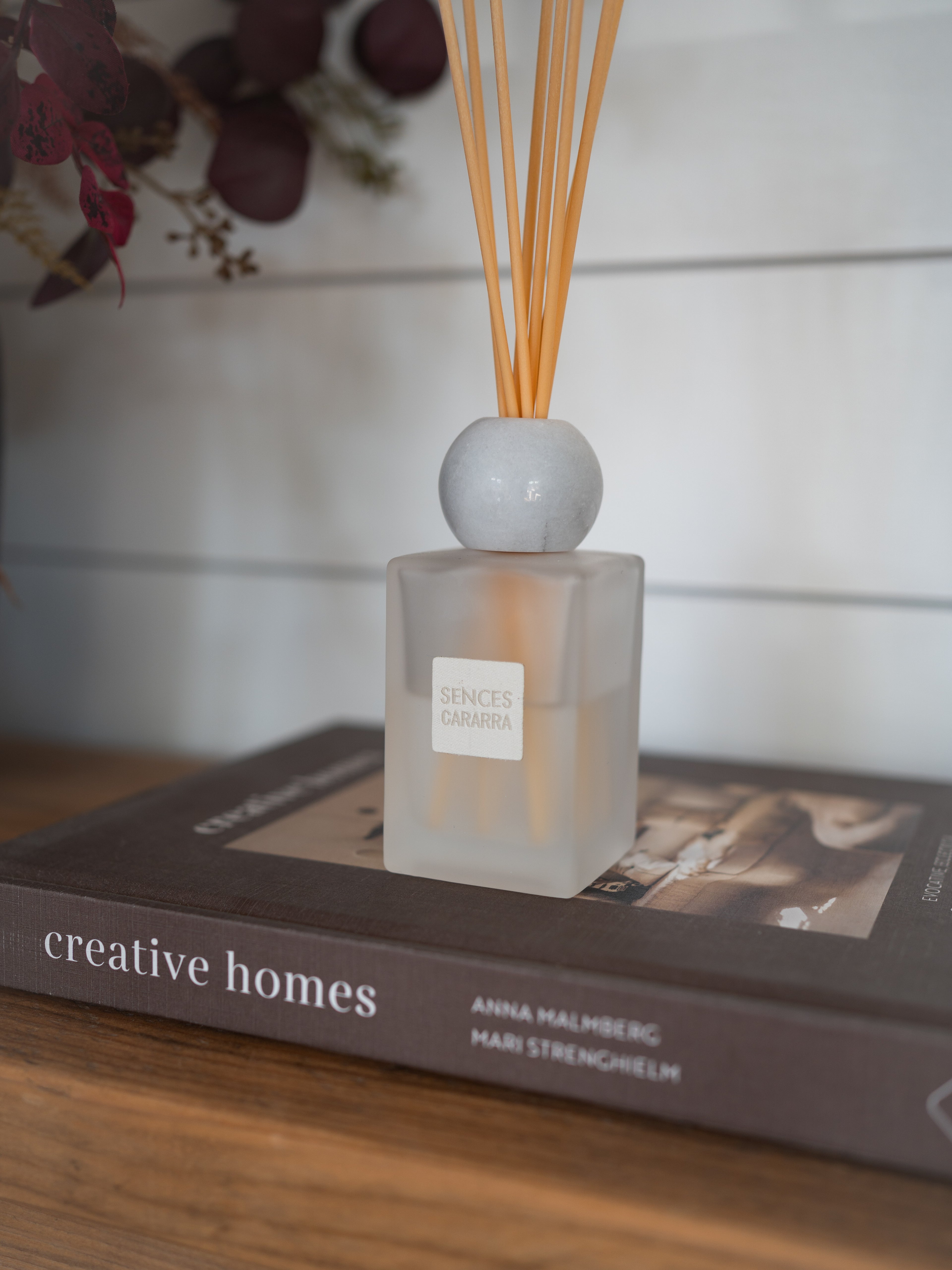 A reed diffuser bottle with a frosted glass body, a round white marble stopper, and natural reed sticks, displayed on top of a book in a styled indoor setting.