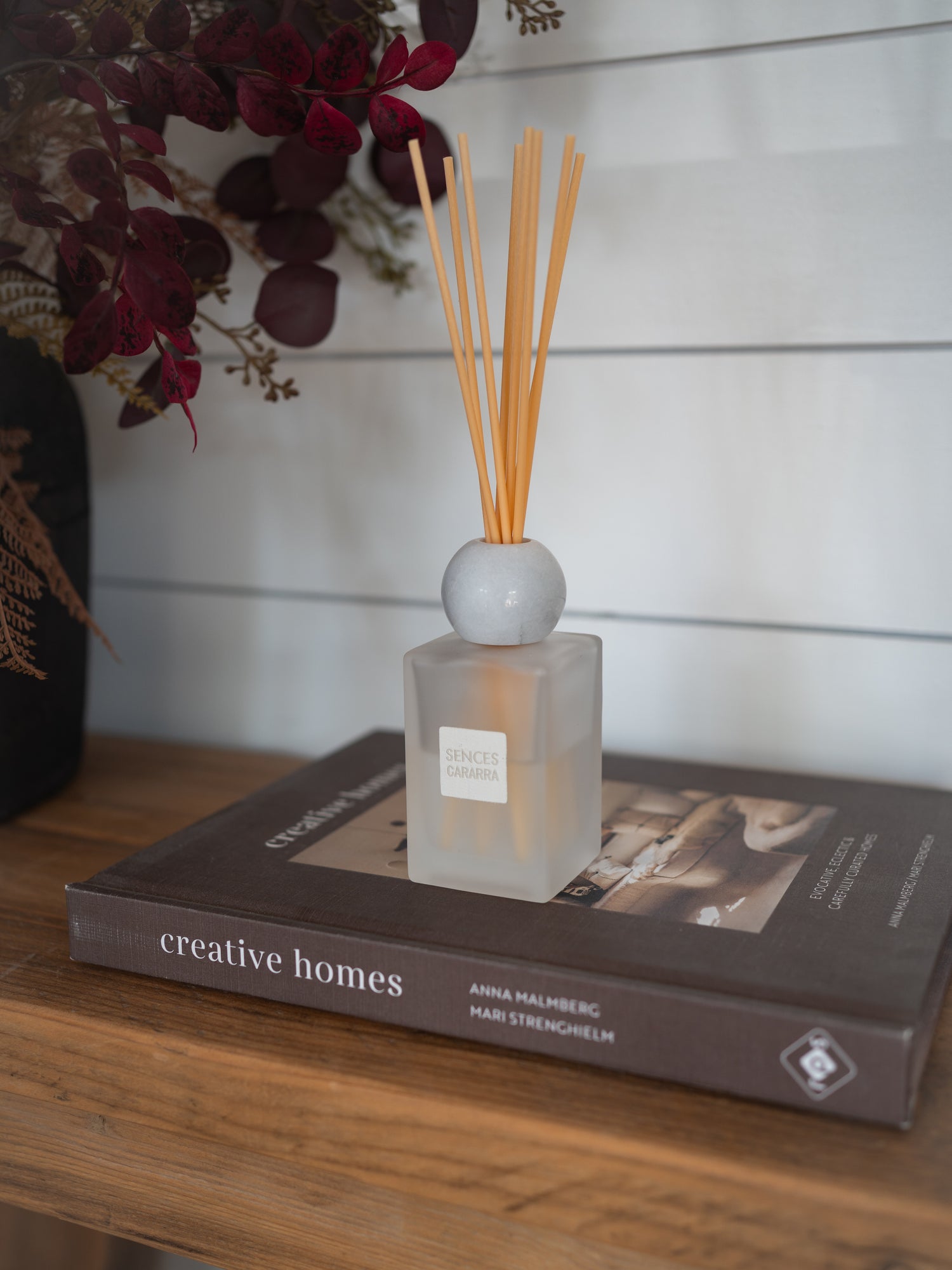 A frosted glass reed diffuser bottle with light wood reeds and a white spherical marble top, placed on a dark book on a wooden surface against a white shiplap wall.