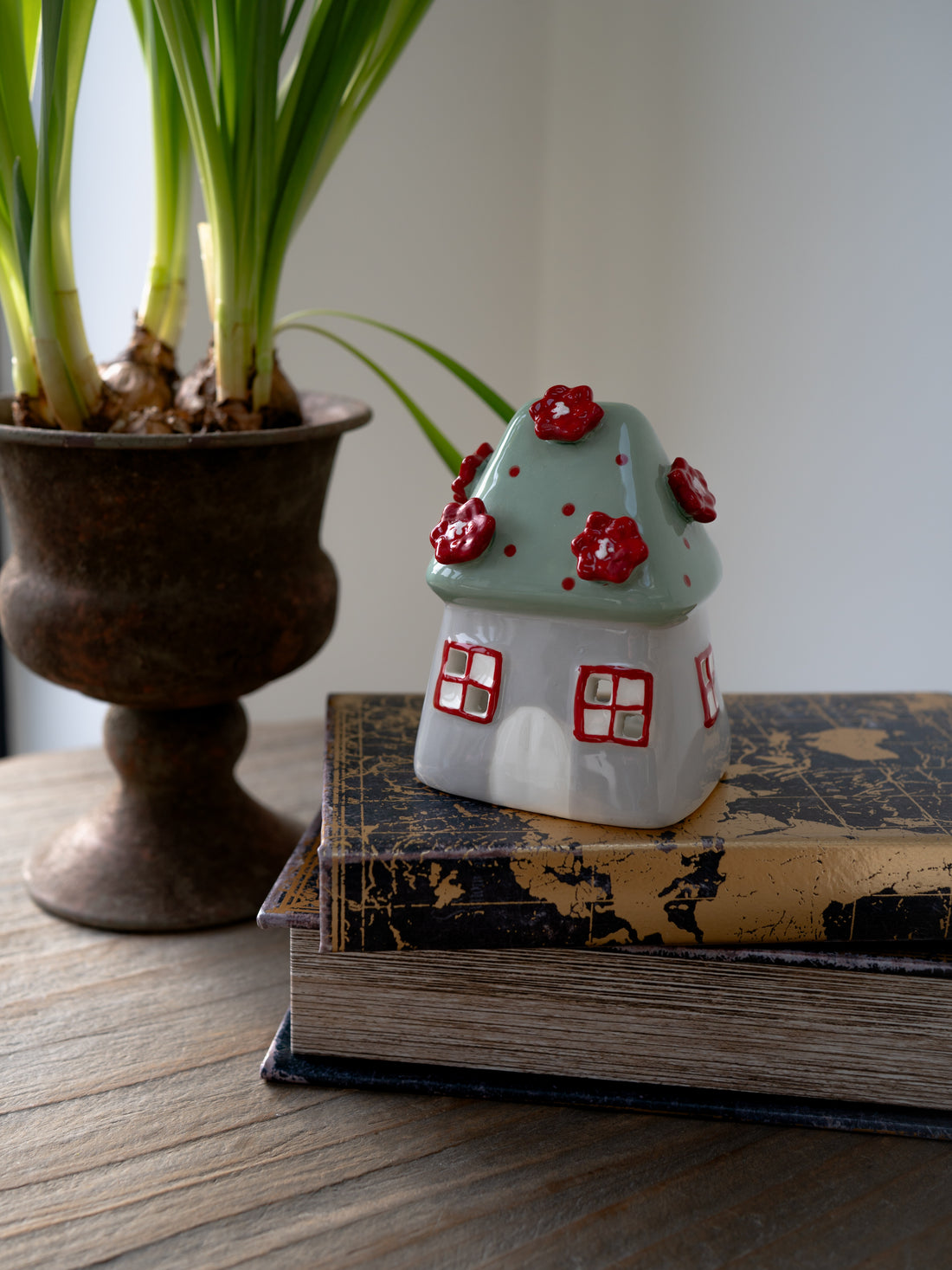 Small ceramic house with red flowers on a stack of books next to a potted plant.