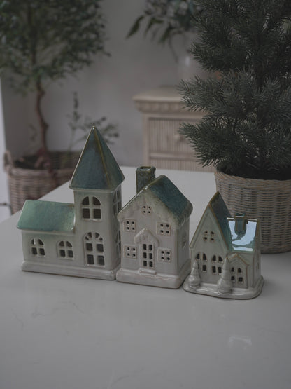 Set of three ceramic house models on a white surface with plants in the background