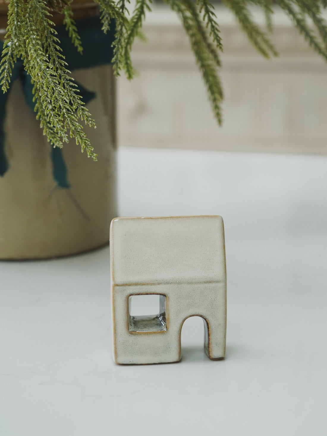 Small ceramic house-shaped object on a light surface with a plant in the background