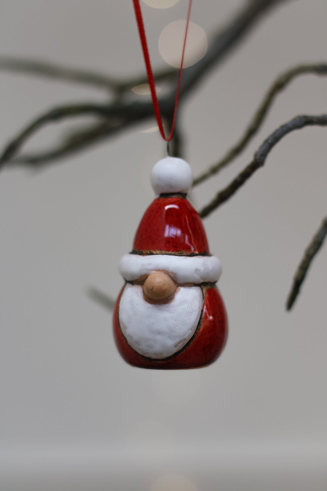 A small Santa Claus christmas tree ornament hanging from a red string, with a glossy red hat and beard, white trim, and a wooden nose, photographed against a soft blurred background with tree branches.