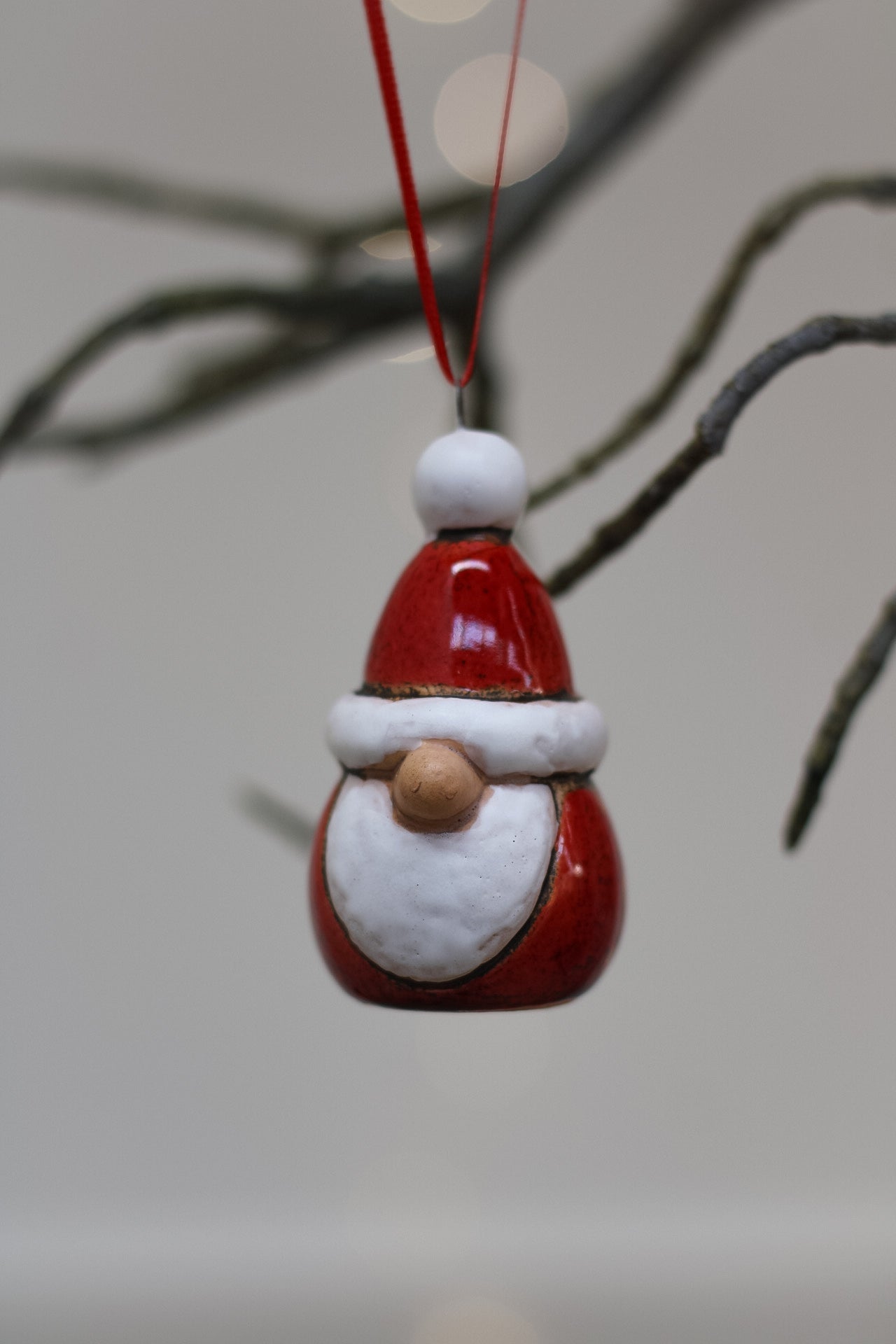 A small Santa Claus christmas tree ornament hanging from a red string, with a glossy red hat and beard, white trim, and a wooden nose, photographed against a soft blurred background with tree branches.