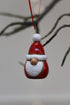 A small Santa Claus christmas tree ornament hanging from a red string, with a glossy red hat and beard, white trim, and a wooden nose, photographed against a soft blurred background with tree branches.