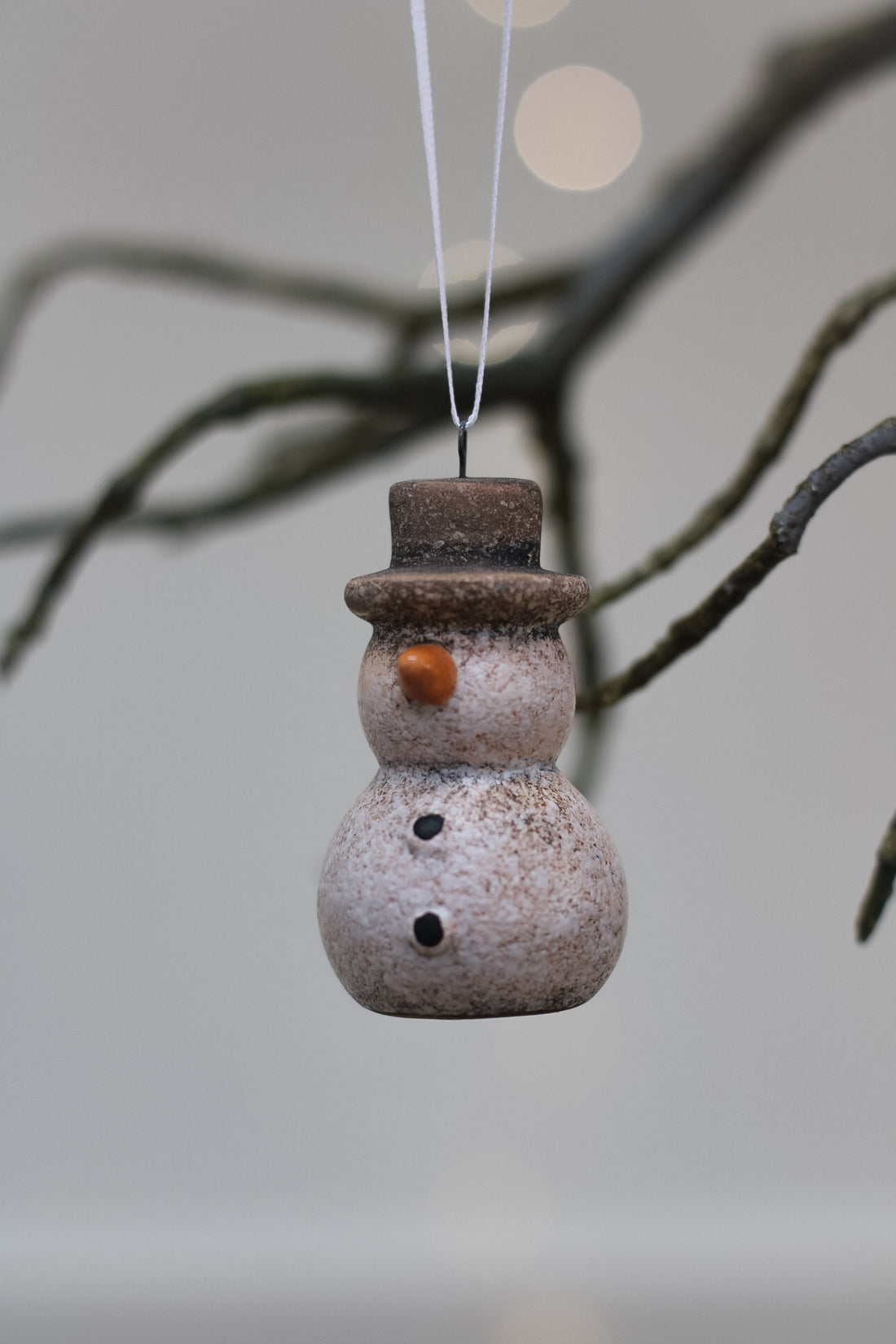 A small ceramic snowman christmas tree ornament hanging from a thin white string, with a speckled light beige body, dark brown hat and buttons, and an orange carrot nose against a softly blurred gray background with tree branches.