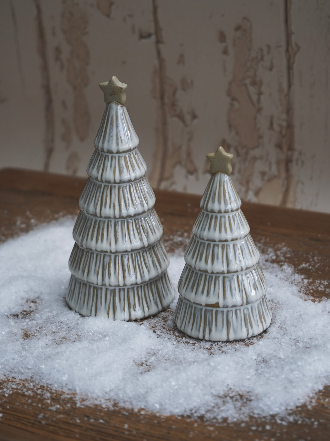 Two ceramic Christmas tree figurines with stars on a wooden surface with a snowy effect.