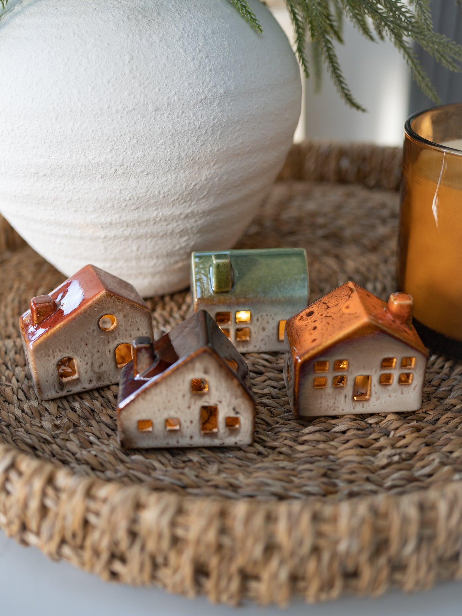Set of small ceramic houses on a woven surface with a white vase and glass container in the background.