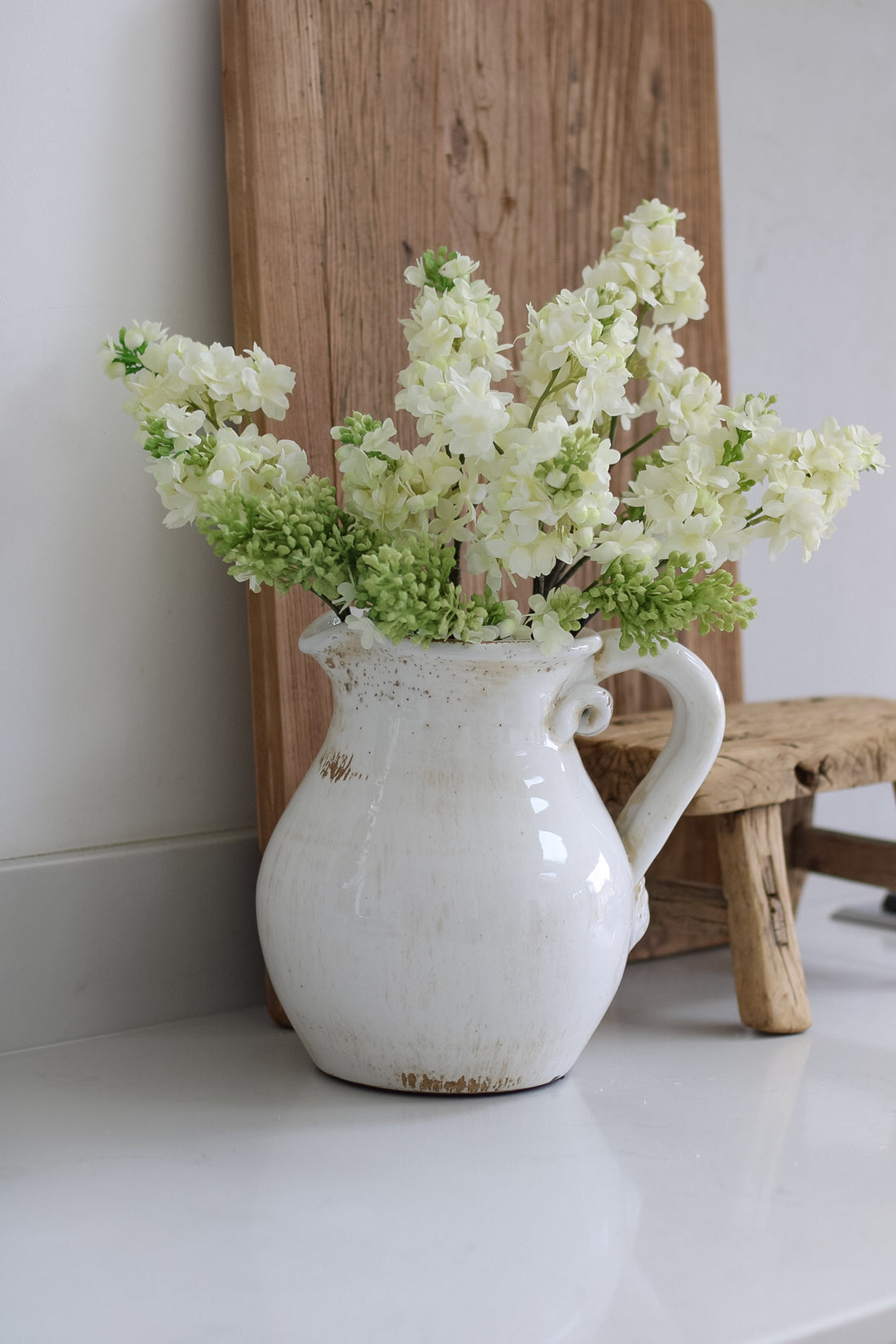 White vase with flowers on a white surface with a wooden board in the background