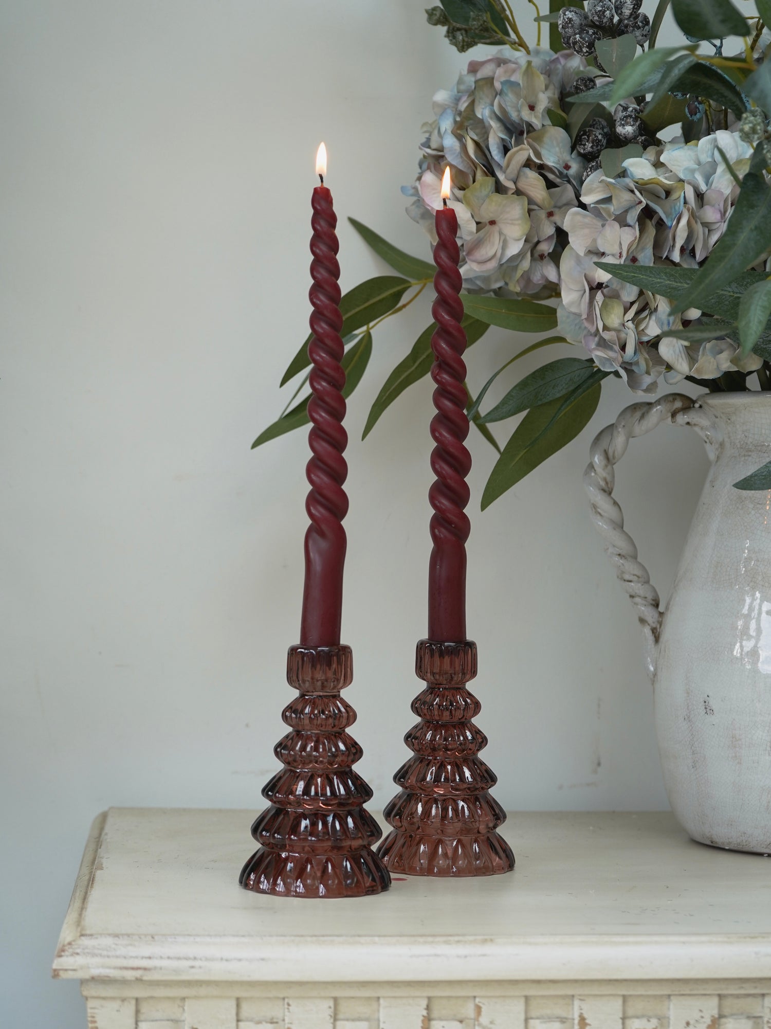 Two twisted red candles in cherry glass christmas tree candle holders on a white surface with a decorative vase in the background.