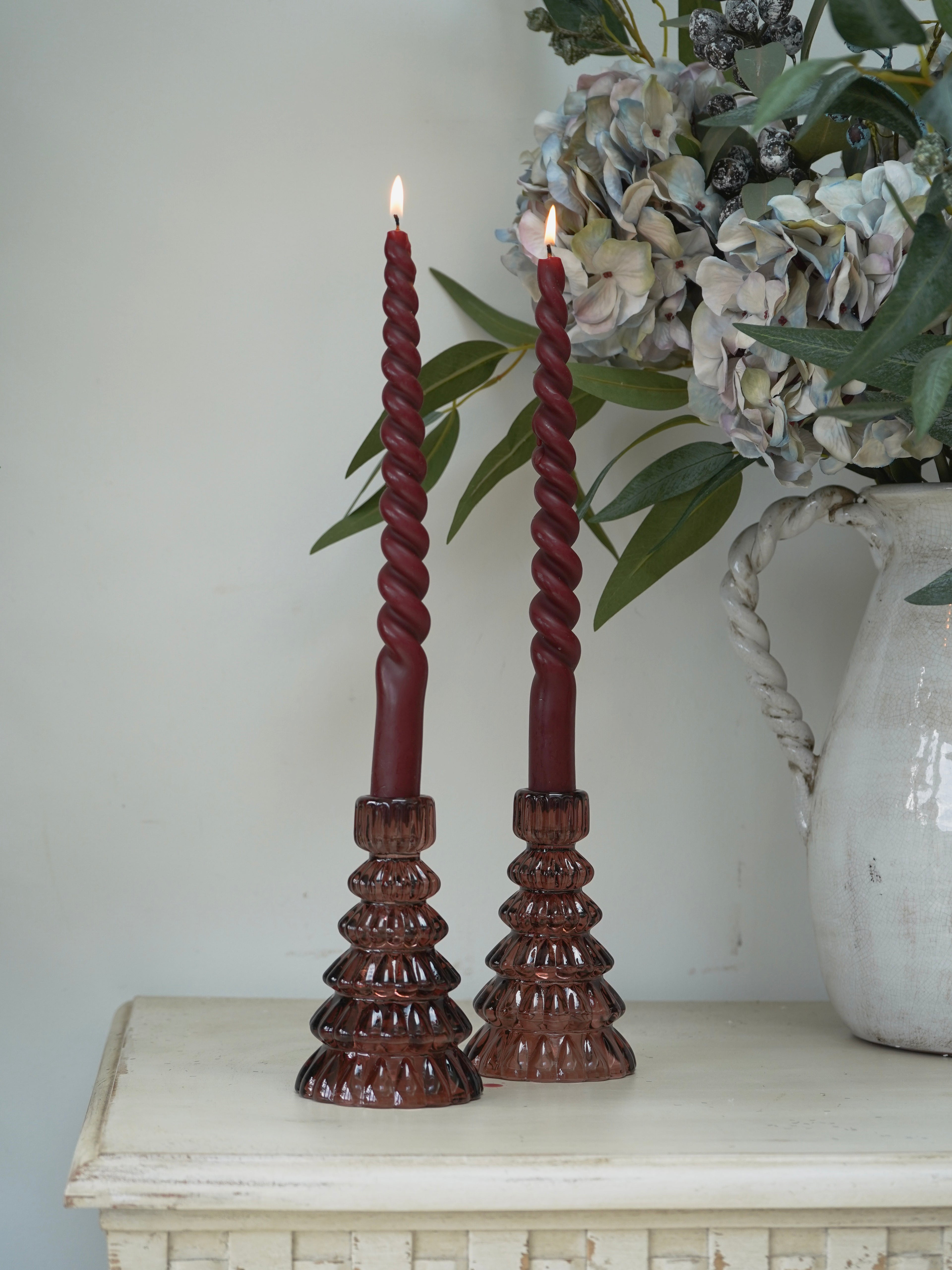 Two twisted red candles in cherry glass christmas tree candle holders on a white surface with a decorative vase in the background.