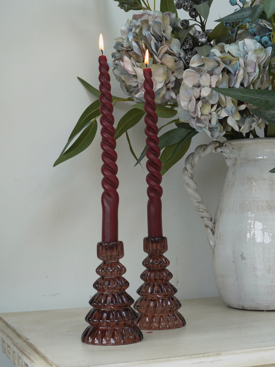 Twisted red candles in christmas tree glass candle holders on a wooden stand with a vase of flowers in the background.