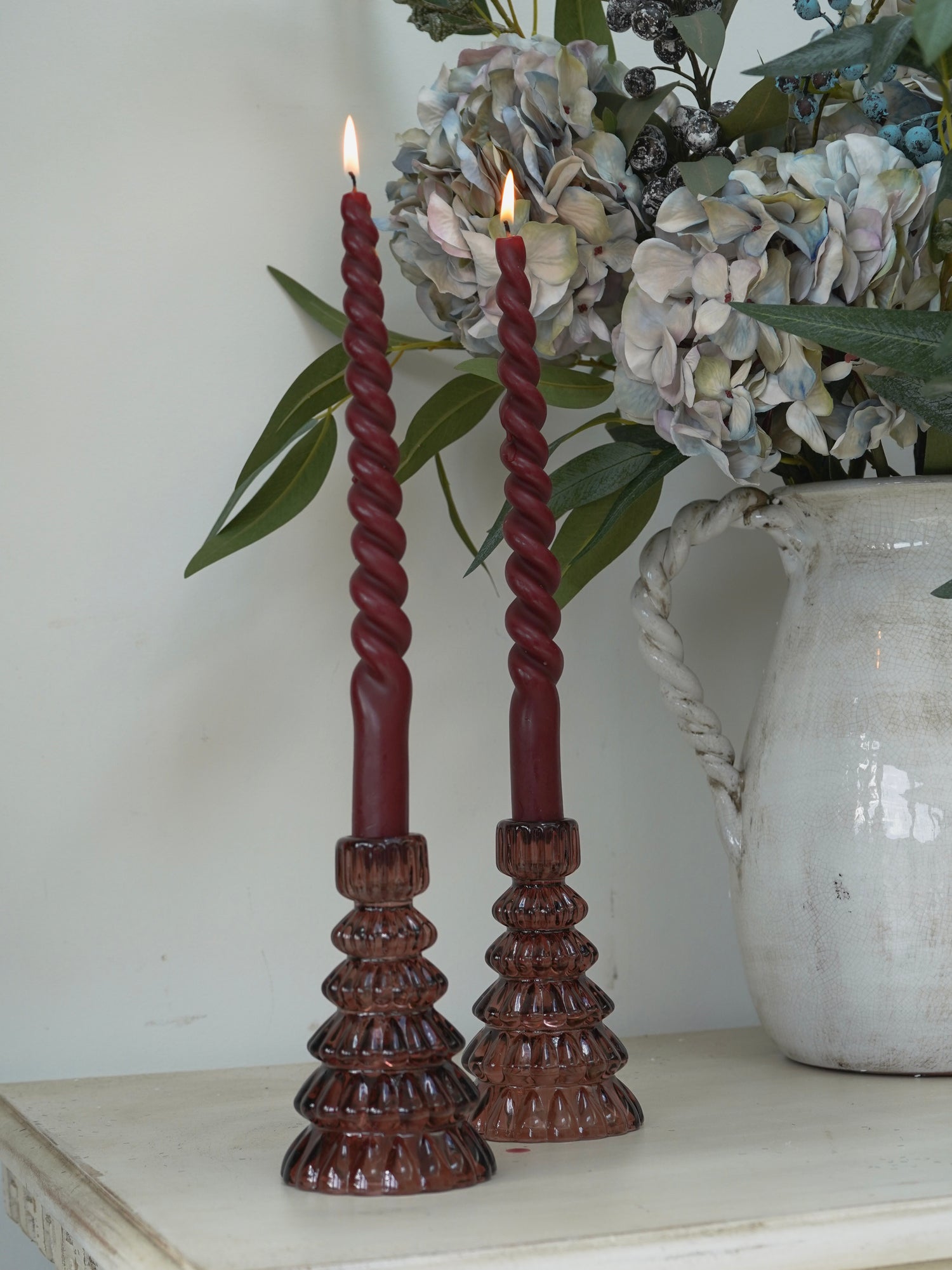 Twisted red candles in christmas tree glass candle holders on a wooden stand with a vase of flowers in the background.