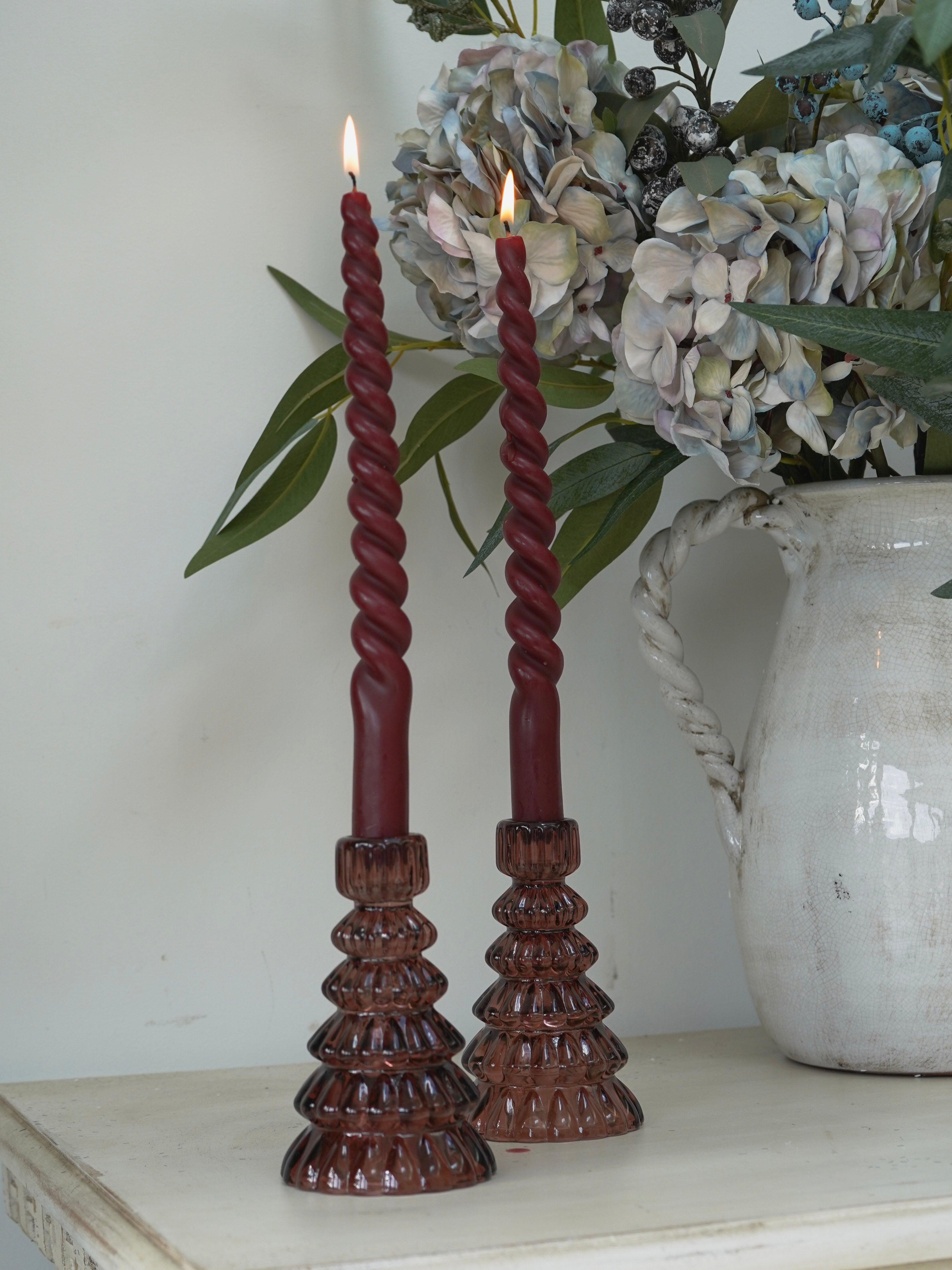 Twisted red candles in christmas tree glass candle holders on a wooden stand with a vase of flowers in the background.