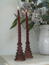 Twisted red candles in christmas tree glass candle holders on a wooden stand with a vase of flowers in the background.