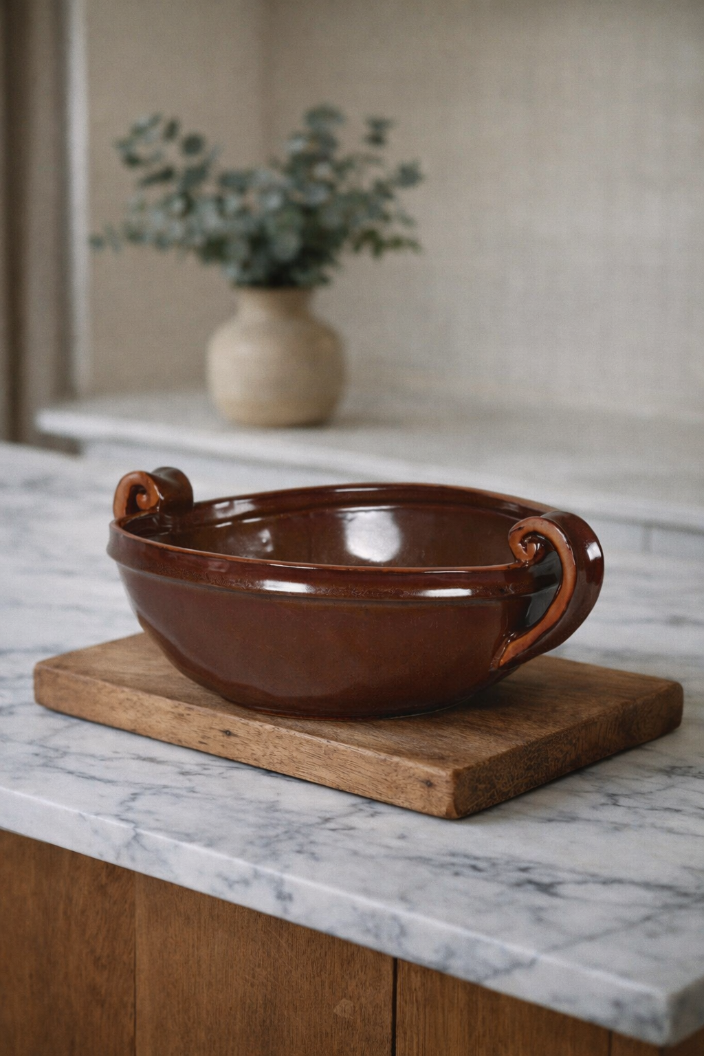 Brown ceramic bowl with handles on a marble surface, with a vase of greenery in the background.