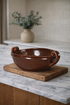 Brown ceramic bowl with handles on a marble surface, with a vase of greenery in the background.