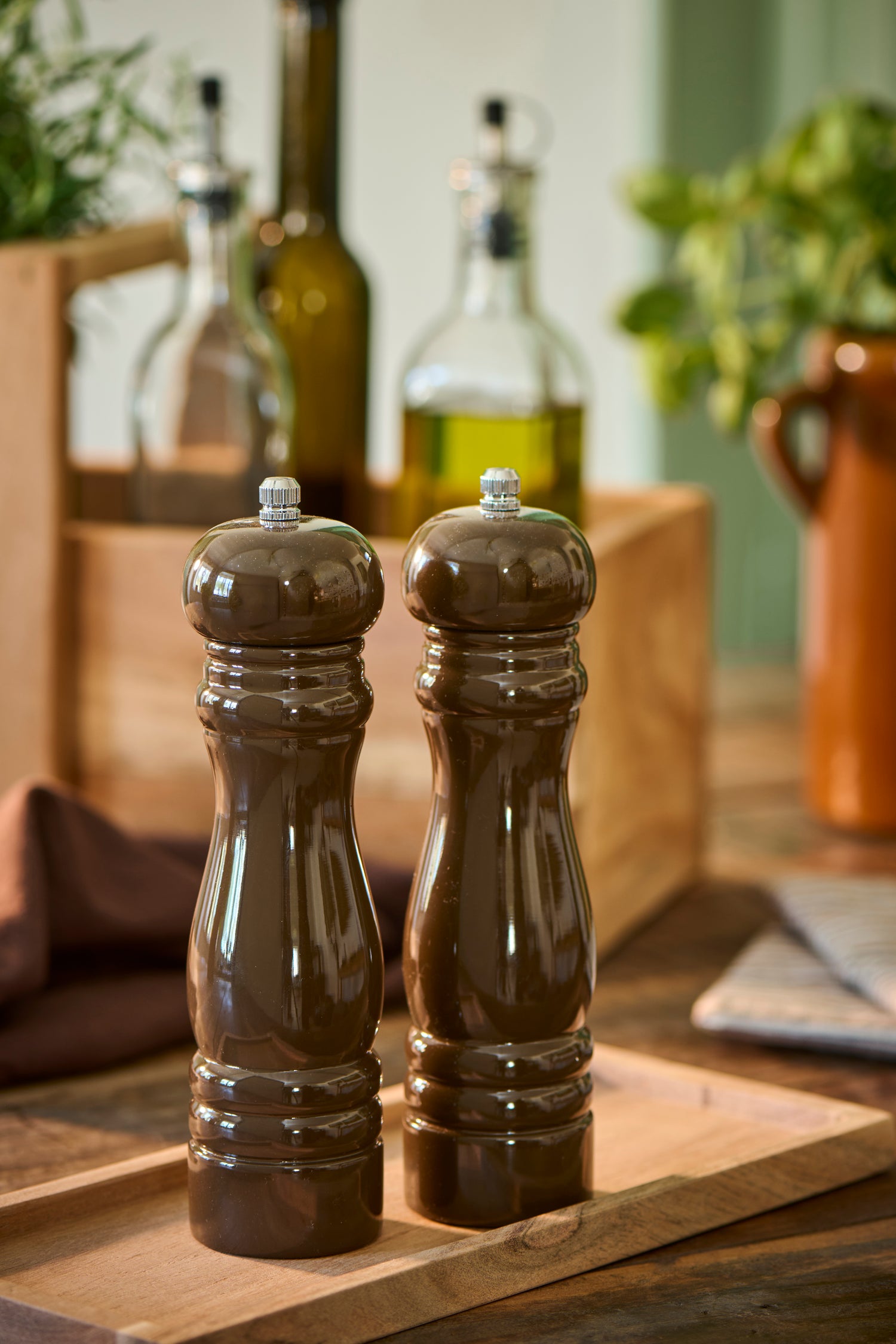 Two dark brown chocolate wooden pepper mills with silver metal tops, standing side-by-side on a wooden cutting board in a kitchen setting.