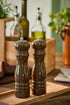 Two dark brown chocolate wooden pepper mills with silver metal tops, standing side-by-side on a wooden cutting board in a kitchen setting.