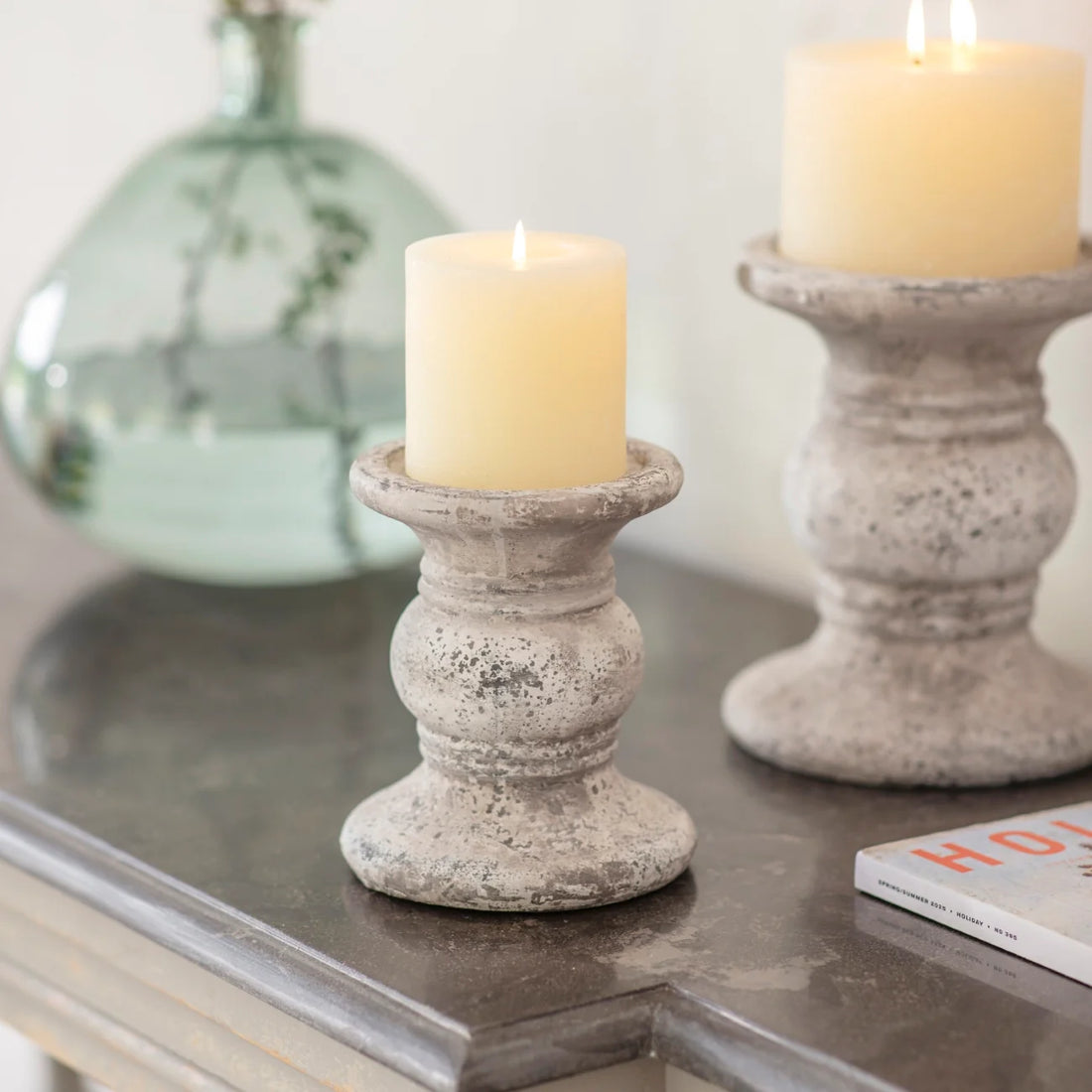 Two cement stoneware candle holders with candles on a reflective surface, one with a blurred vase and magazine in the background.