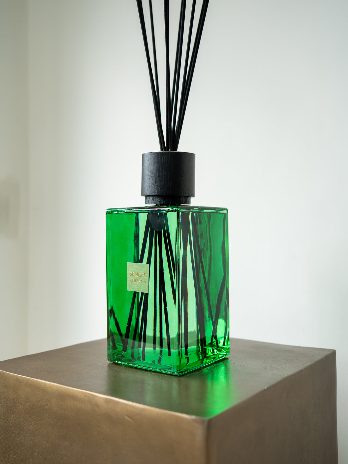 A green glass reed diffuser bottle with black reeds and a black cap, displayed on a metallic pedestal against a light gray background.