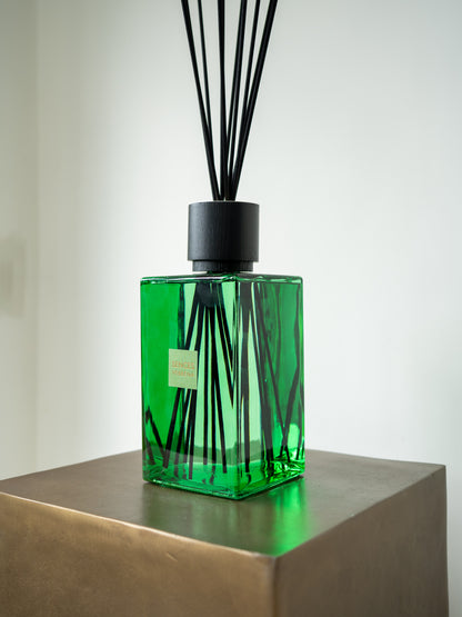 A green glass reed diffuser bottle with black reeds and a black cap, displayed on a metallic pedestal against a light gray background.