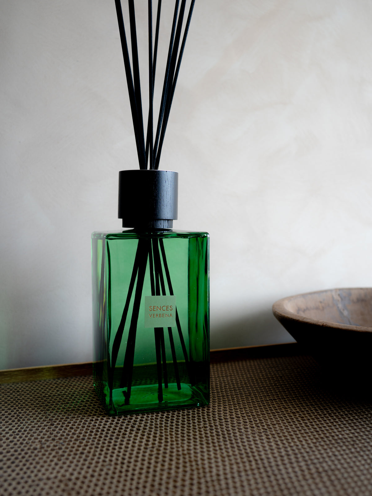 A green glass SENCES CITRUS VERBENA reed diffuser bottle with black reeds and a black cap, standing on a woven surface against a light beige wall. A small label is visible on the bottle.