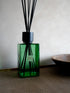 A green glass SENCES CITRUS VERBENA reed diffuser bottle with black reeds and a black cap, standing on a woven surface against a light beige wall. A small label is visible on the bottle.