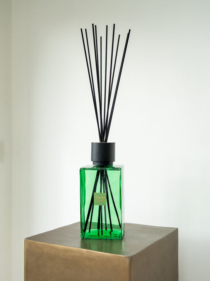 A green glass SENCES CITRUS VERBENA reed diffuser bottle with black diffuser sticks, displayed on a metallic gold pedestal against a plain off-white wall.