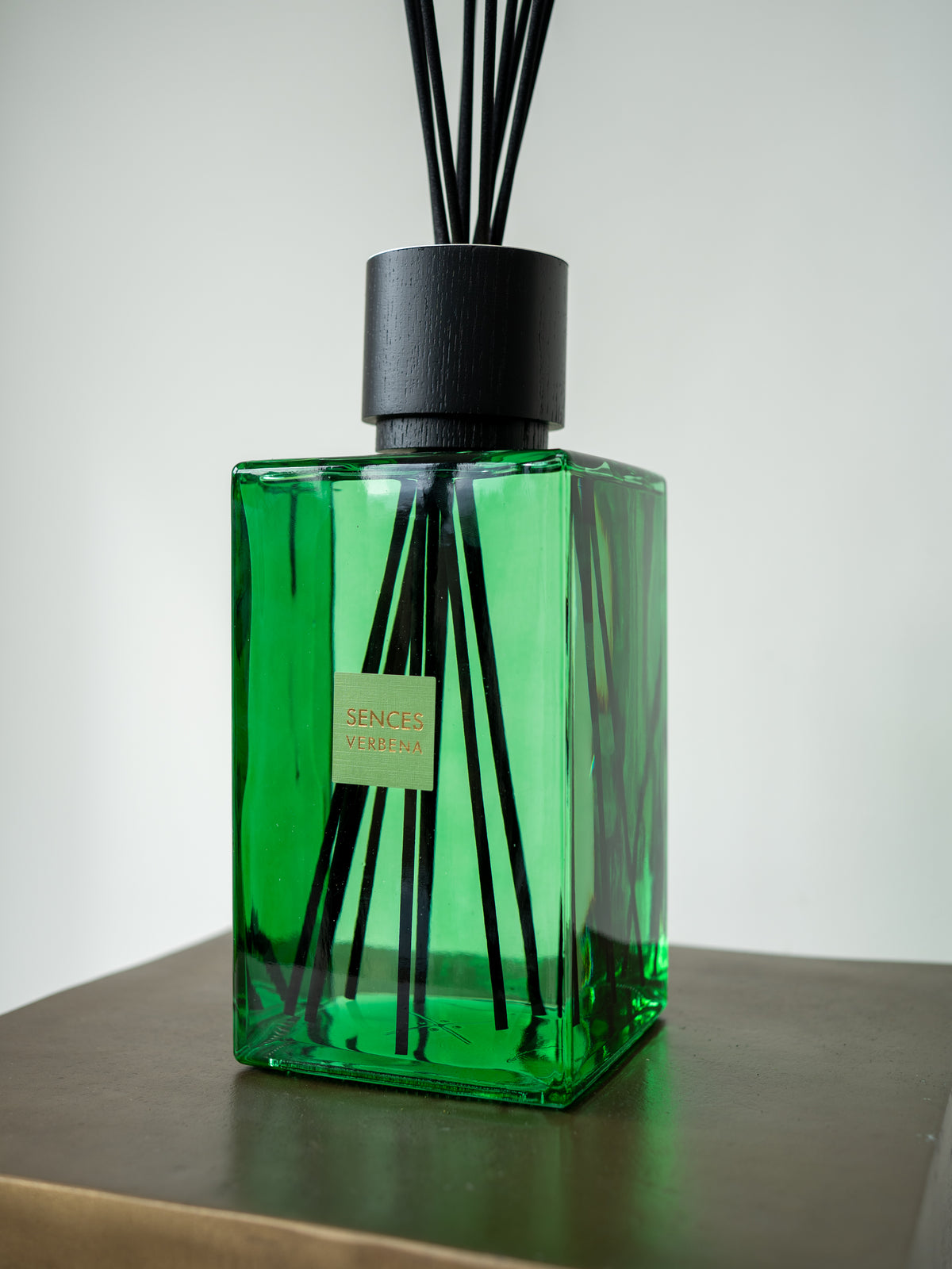 A green glass reed diffuser bottle with black reeds and a black cap, sitting on a dark wooden surface against a light gray background.