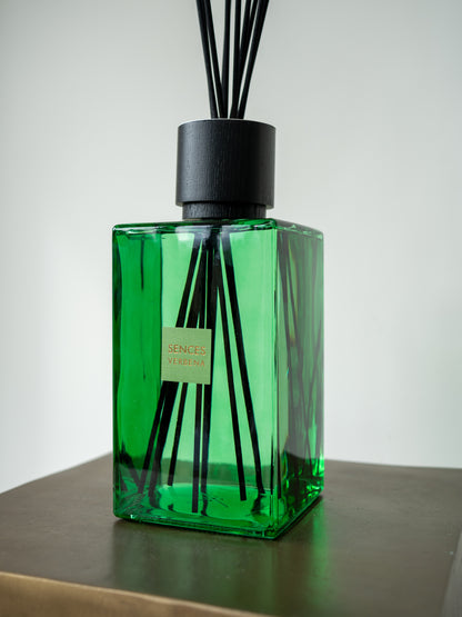 A green glass reed diffuser bottle with black reeds and a black cap, sitting on a dark wooden surface against a light gray background.