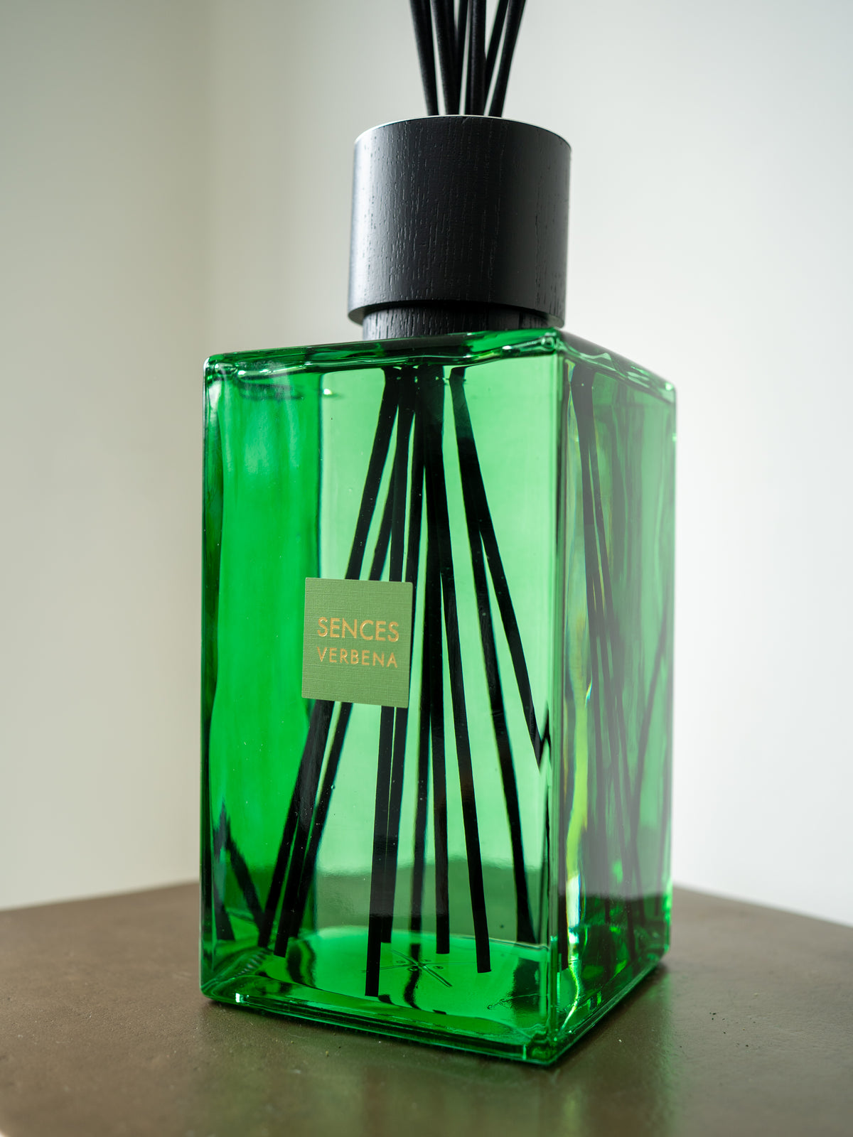 A green glass reed diffuser bottle with black reeds and a black cap, sitting on a dark brown surface against a light gray background. A small label on the front reads &