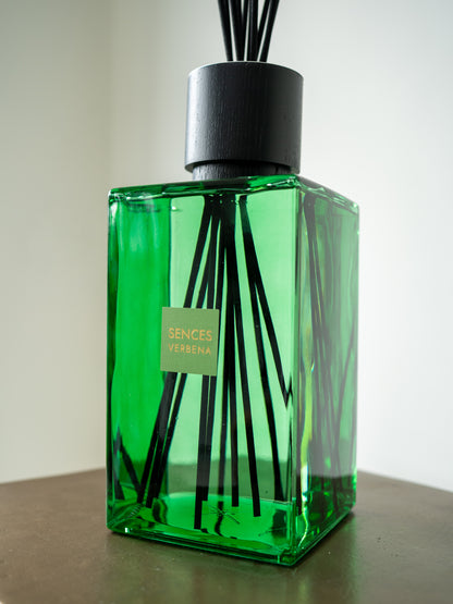A green glass reed diffuser bottle with black reeds and a black cap, sitting on a dark brown surface against a light gray background. A small label on the front reads &