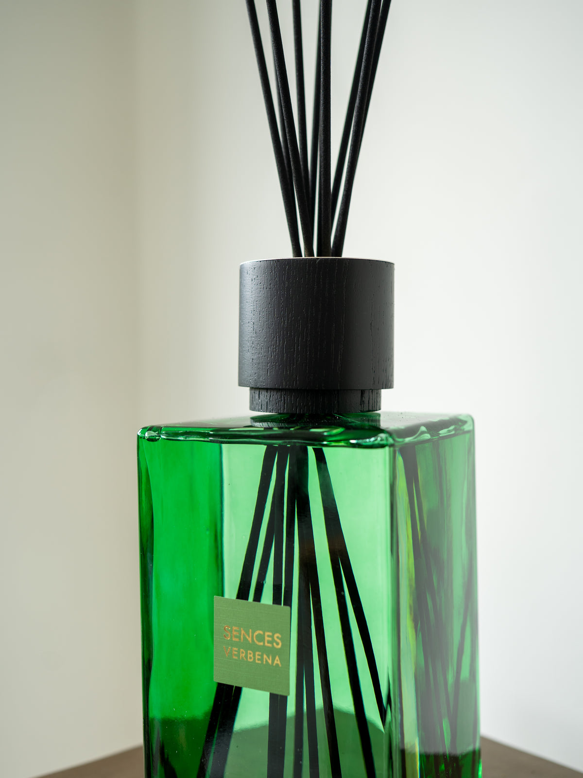 A green glass reed diffuser bottle with black reeds and a black cap, featuring a green label that reads &