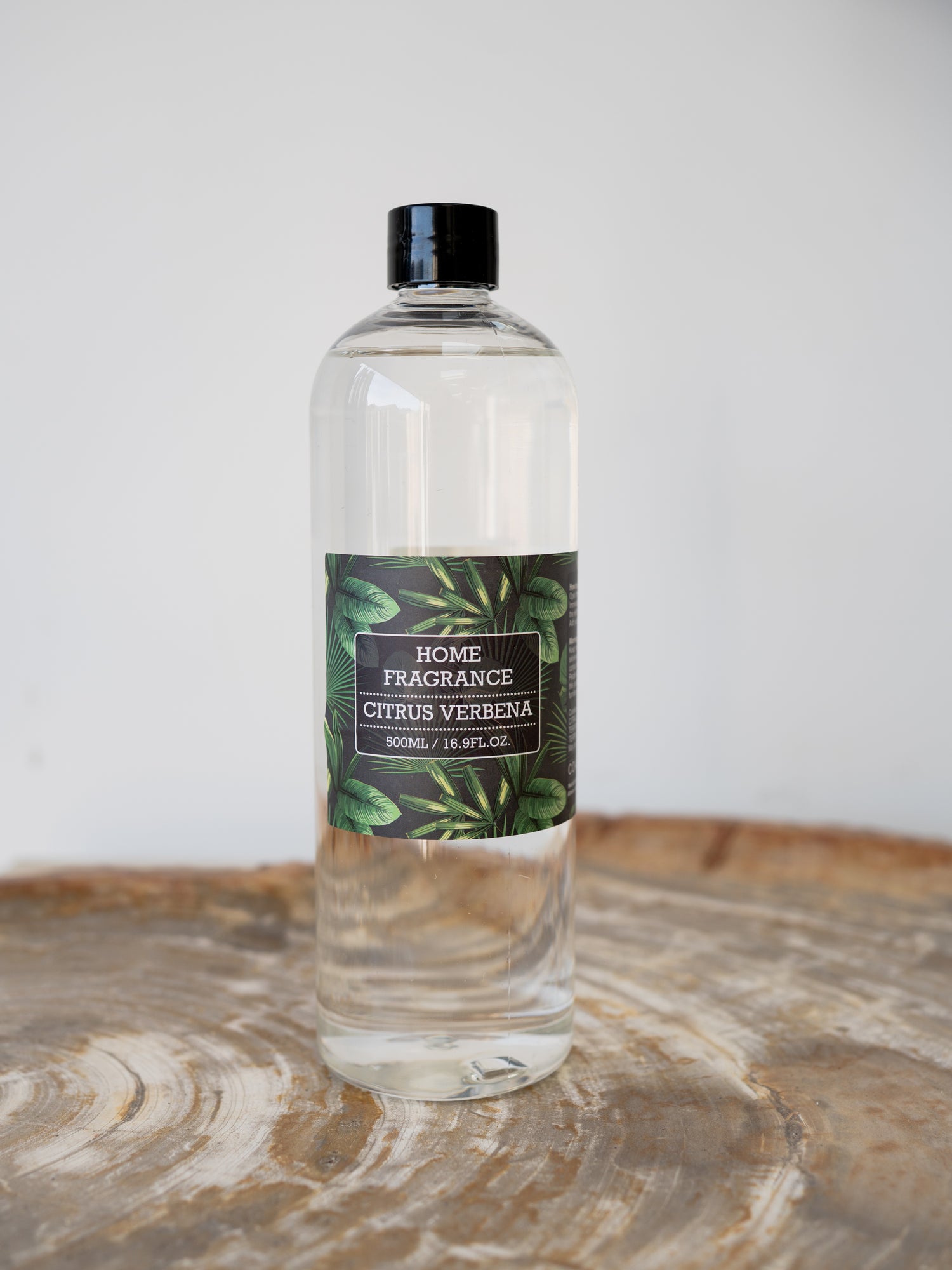 A clear bottle of Home Fragrance Citrus Verbena home fragrance diffuser refill oil with a black cap and a dark green botanical label, standing on a rustic wood slice against a light gray background.