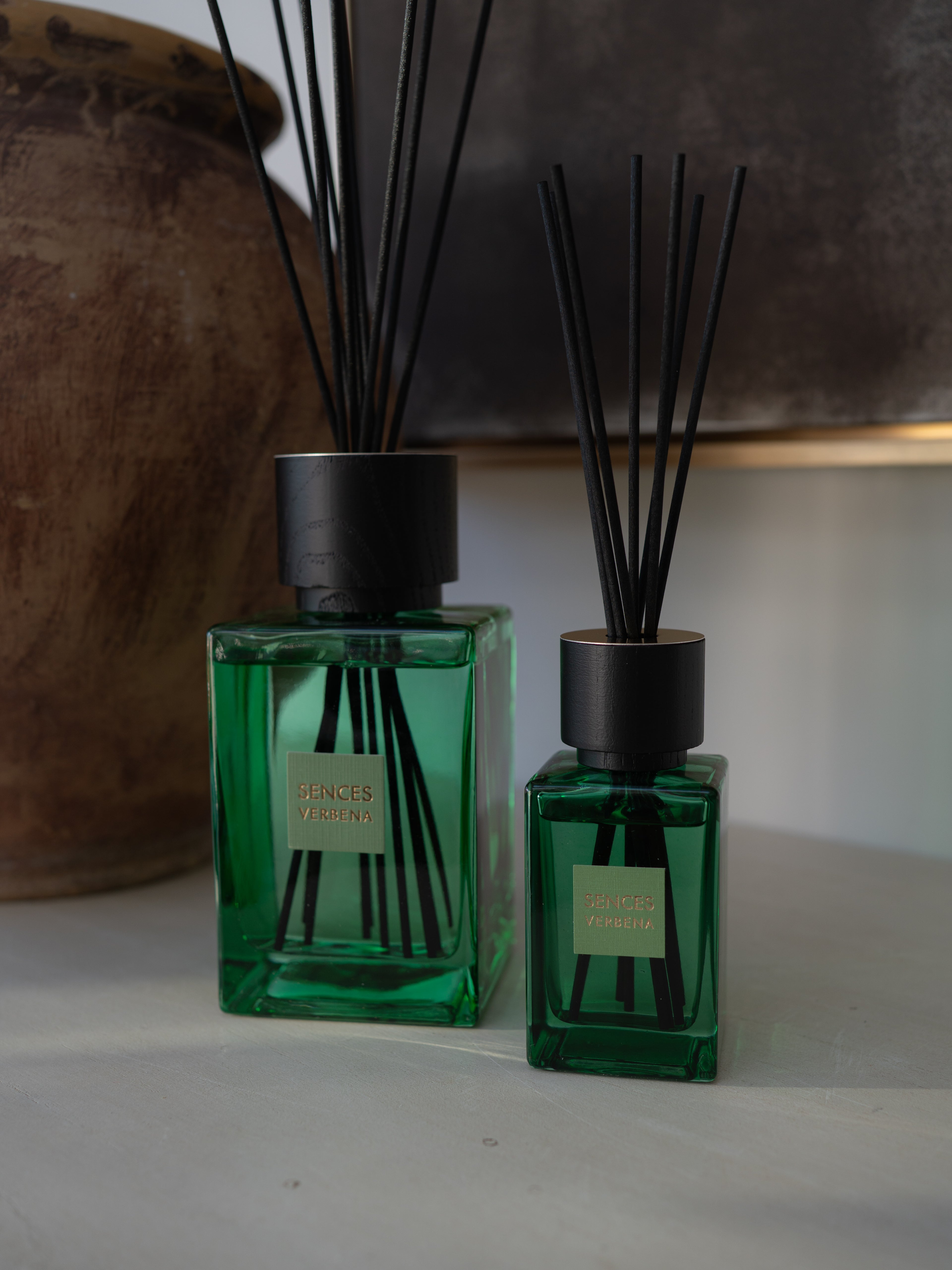 Two green glass reed diffusers with black caps and black reeds, standing on a light surface with a blurred brown ceramic vessel in the background.