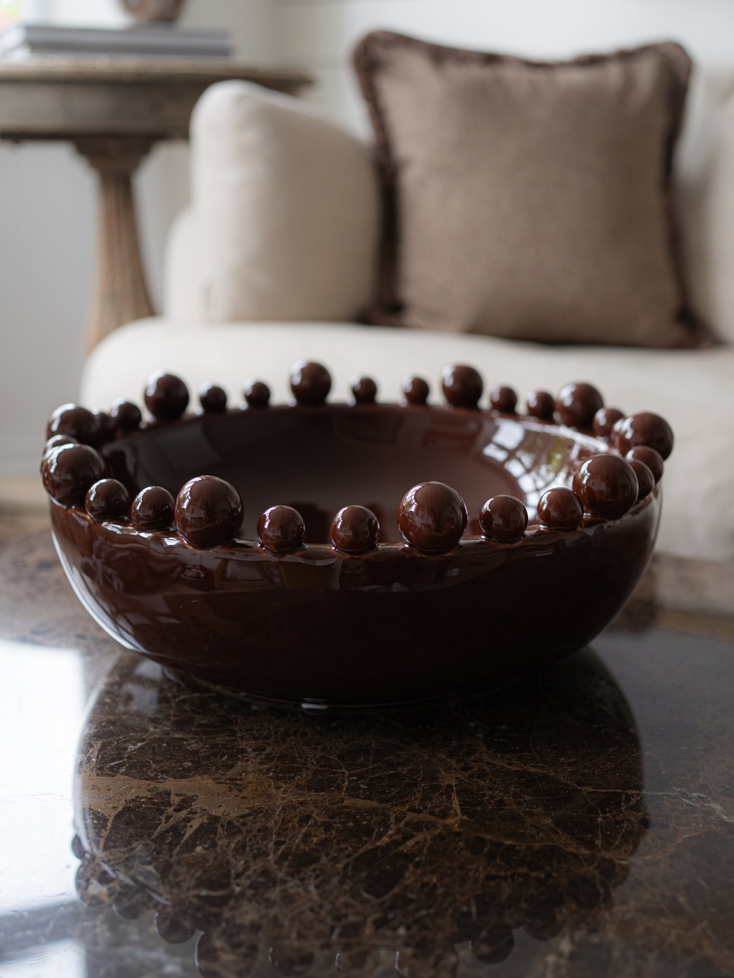 A dark brown ceramic bowl with a raised beaded bobbled rim, displayed on a polished dark marble table in a softly lit living room setting.