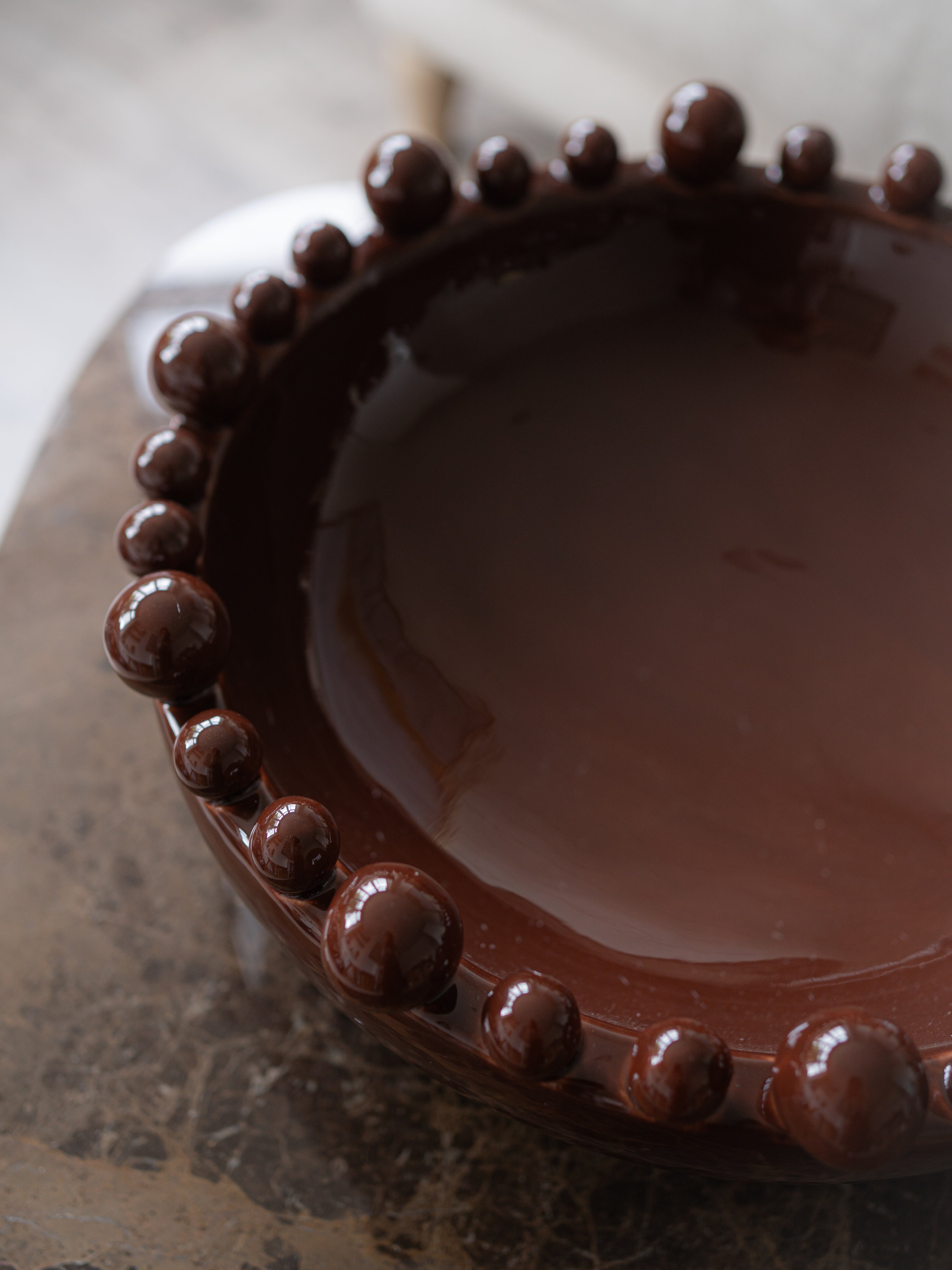 A close-up of a glossy dark brown ceramic bowl with a raised beaded  bobbled rim, resting on a dark stone surface.
