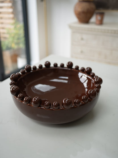 A glossy dark brown ceramic bowl with a raised beaded rim, set on a light-colored tabletop in a softly blurred indoor setting.